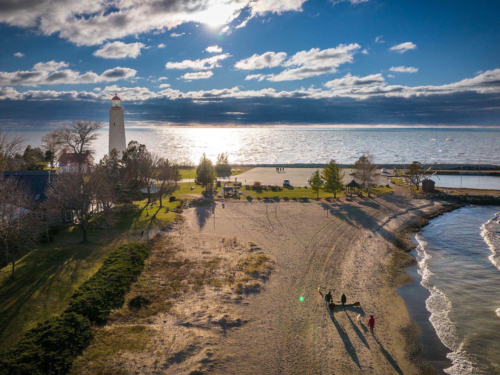 Aerial view of a Lake Huron shoreline park, concrete breakwater arms and a white lighthouse under broken cloud